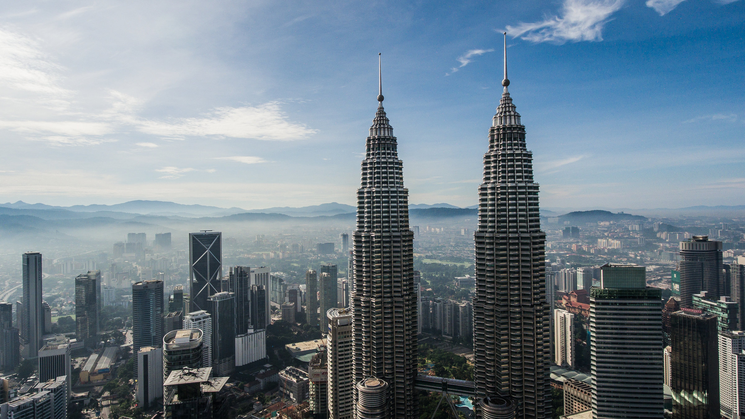"Malaysia von oben": Luftbild-(Schrägbild- ) Aufnahme der Stad Kuala Lumpur. Im Hintergrund mittelhohe Bergketten im Dunst, Stadtlandschaft mit Hochhäusern, im Vordergrund das obere Drittel zweier baugleicher, sich nach oben verjüngender Hochhäuser (Petronas Towers)