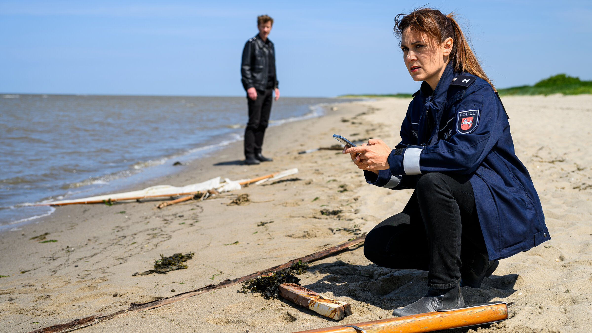 "Friesland – Sturmmöwen": Süher Özlügül (Sophie Dal) kniet am Strand vor zerbrochenen und verstreuten Teilen eines Segelbootes. Im Hintergrund steht Henk Cassens (Maxim Mehmet).
