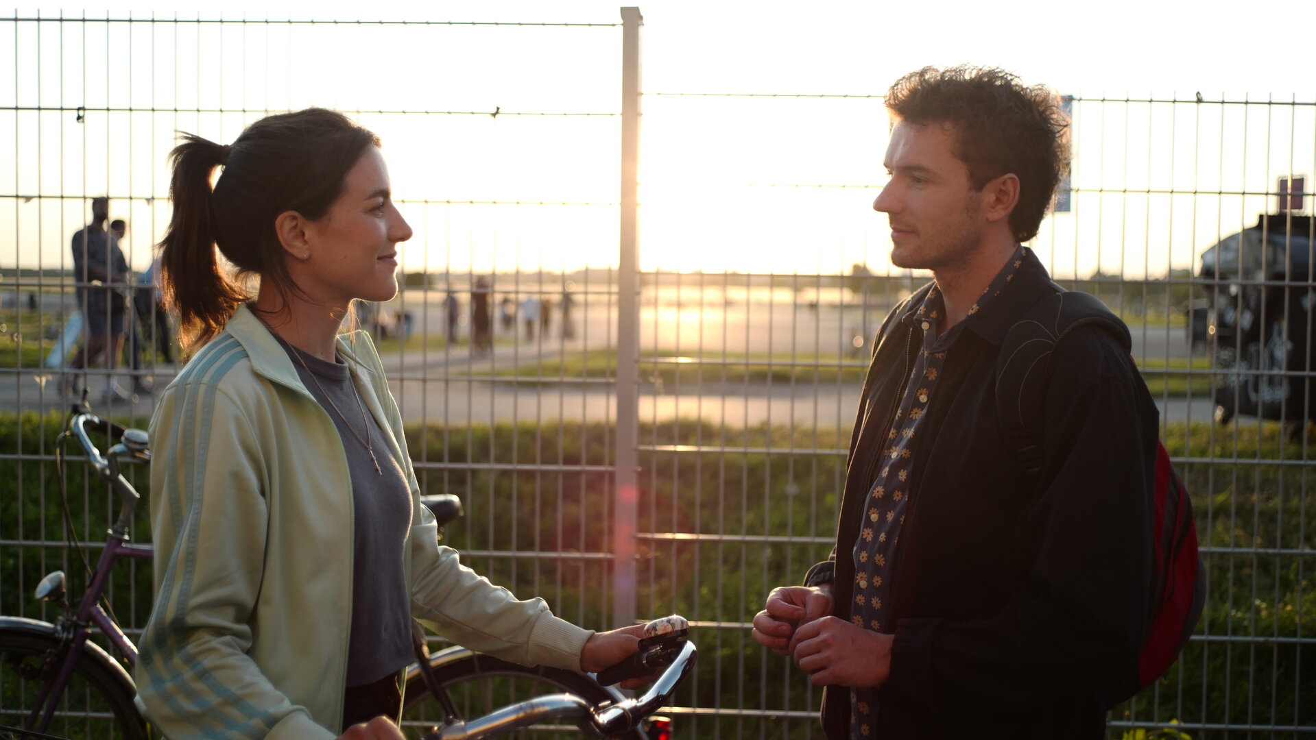 "This Is Gonna Be Great - Dating": Am Tempelhofer Feld: Hanna (Mona Vojacek Koper) und David (Rein Mulder) stehen sich vor einem Zaun gegenüber. Hanna hat die Hände an ihrem Fahrradlenker. Im Hintergrund beginnt der Sonnenuntergang.