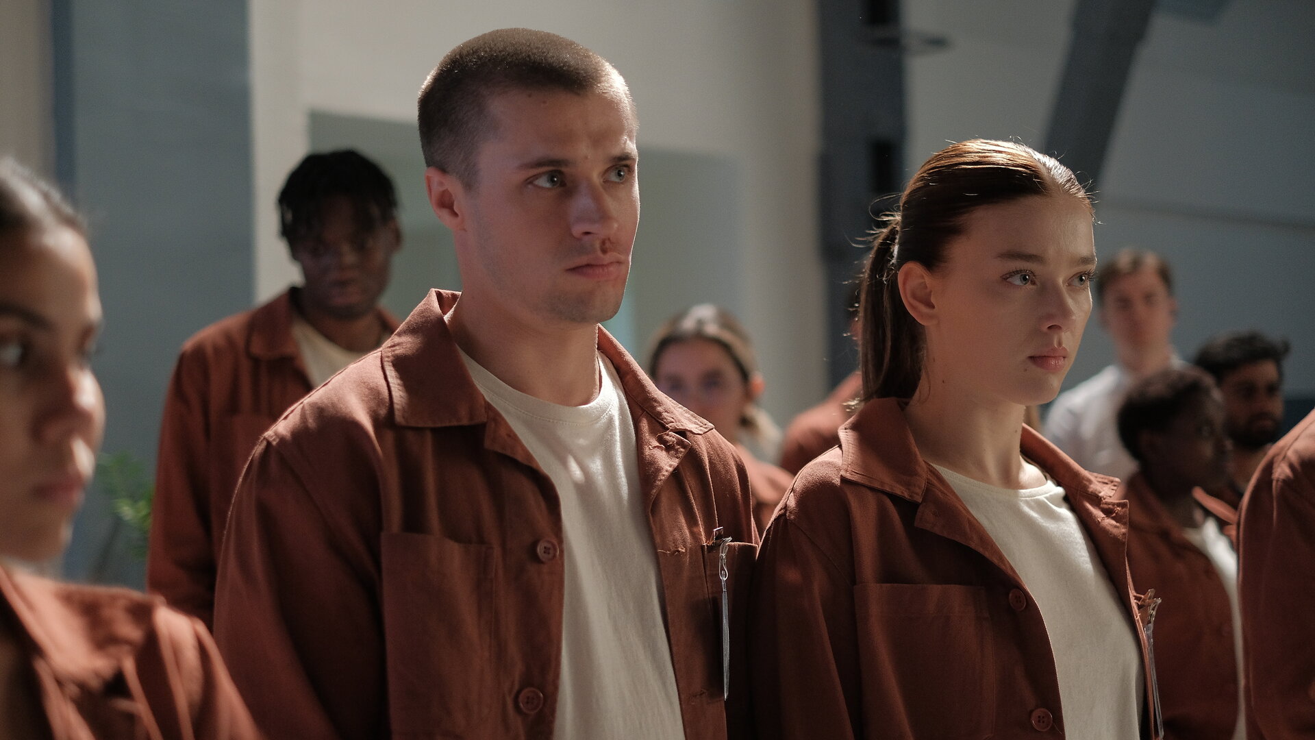 "Fallen - Zusammenhalt": Luce (Jessica Alexander) und Daniel (Gijs Blom) stehen nebeneinander in einer Gruppe von Personen (Komparsen), alle tragen orangene Anzüge mit Namensschildern. Beide blicken ernst geradeaus. Im Hintergrund sind karge, weiße Wände mit einem Stahlpfeiler zu erkennen.