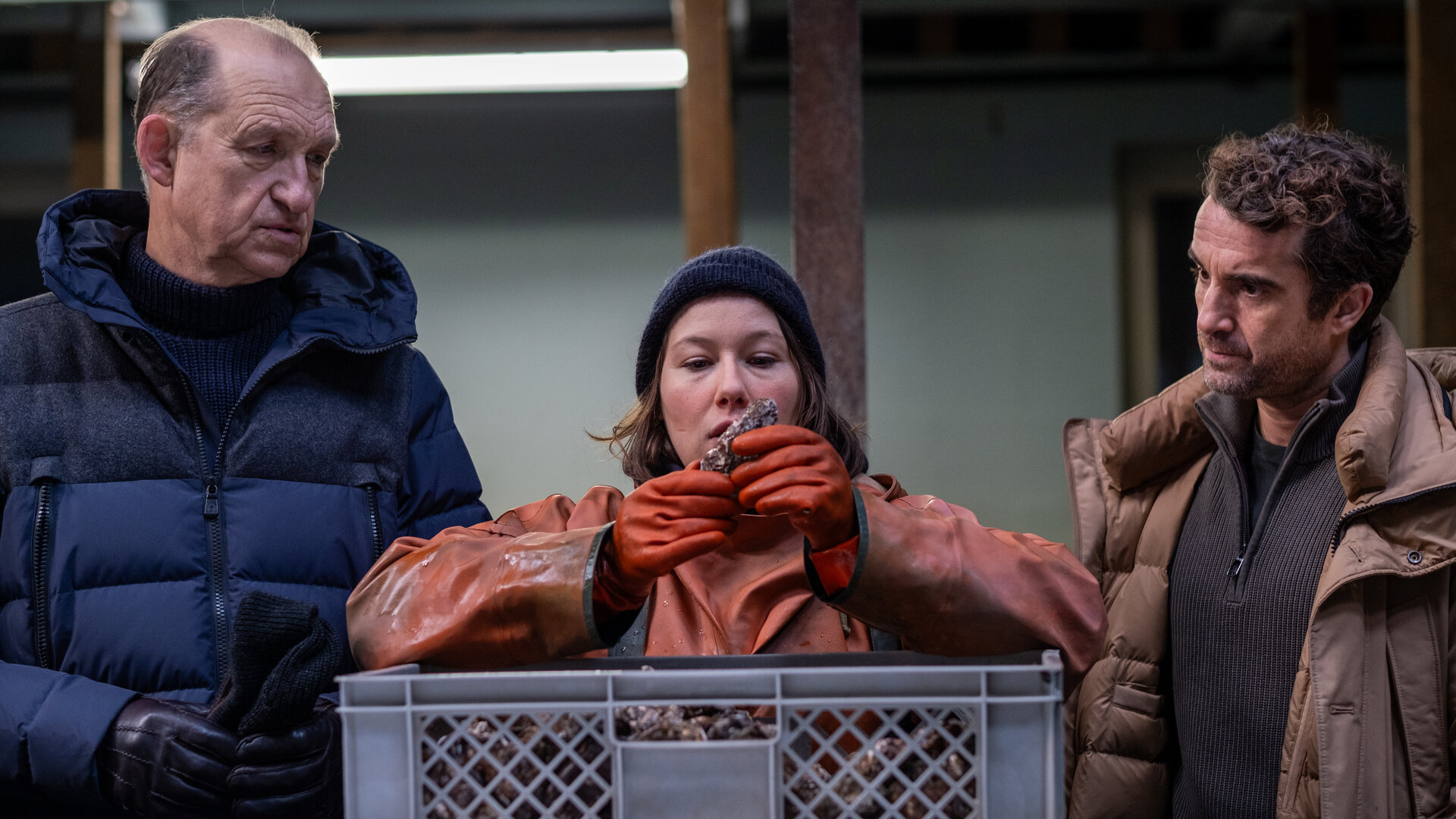 "Nord Nord Mord - Sievers und die stillen Austern": Carl Sievers (Peter Heinrich Brix) und Hinnerk Feldmann (Oliver Wnuk) haben Tomke Bundis (Josefine Isreal) in ihrer Mitte, die gerade mit dicken roten Handschuhen Austern aus einer grauen Lieferkiste fischt und prüft.