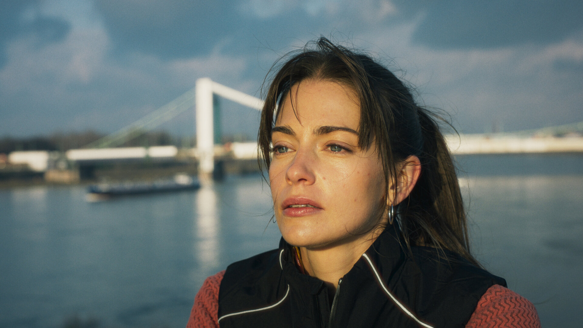 "Bauchgefühl - Lena": Lena (Laura Berlin) steht hoch auf einer Brücke. Im Hintergrund ist eine weitere Brücke zu sehen, unter ihr fährt ein Frachtschiff auf dem breiten Fluss entlang.