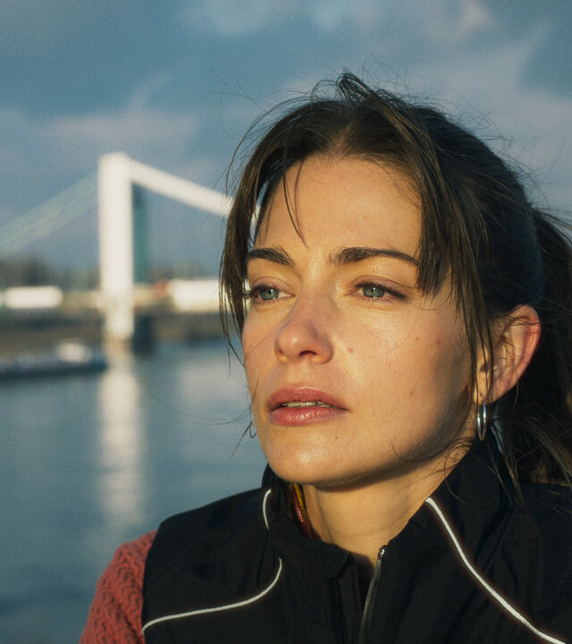 "Bauchgefühl - Lena": Lena (Laura Berlin) steht hoch auf einer Brücke. Im Hintergrund ist eine weitere Brücke zu sehen, unter ihr fährt ein Frachtschiff auf dem breiten Fluss entlang.
