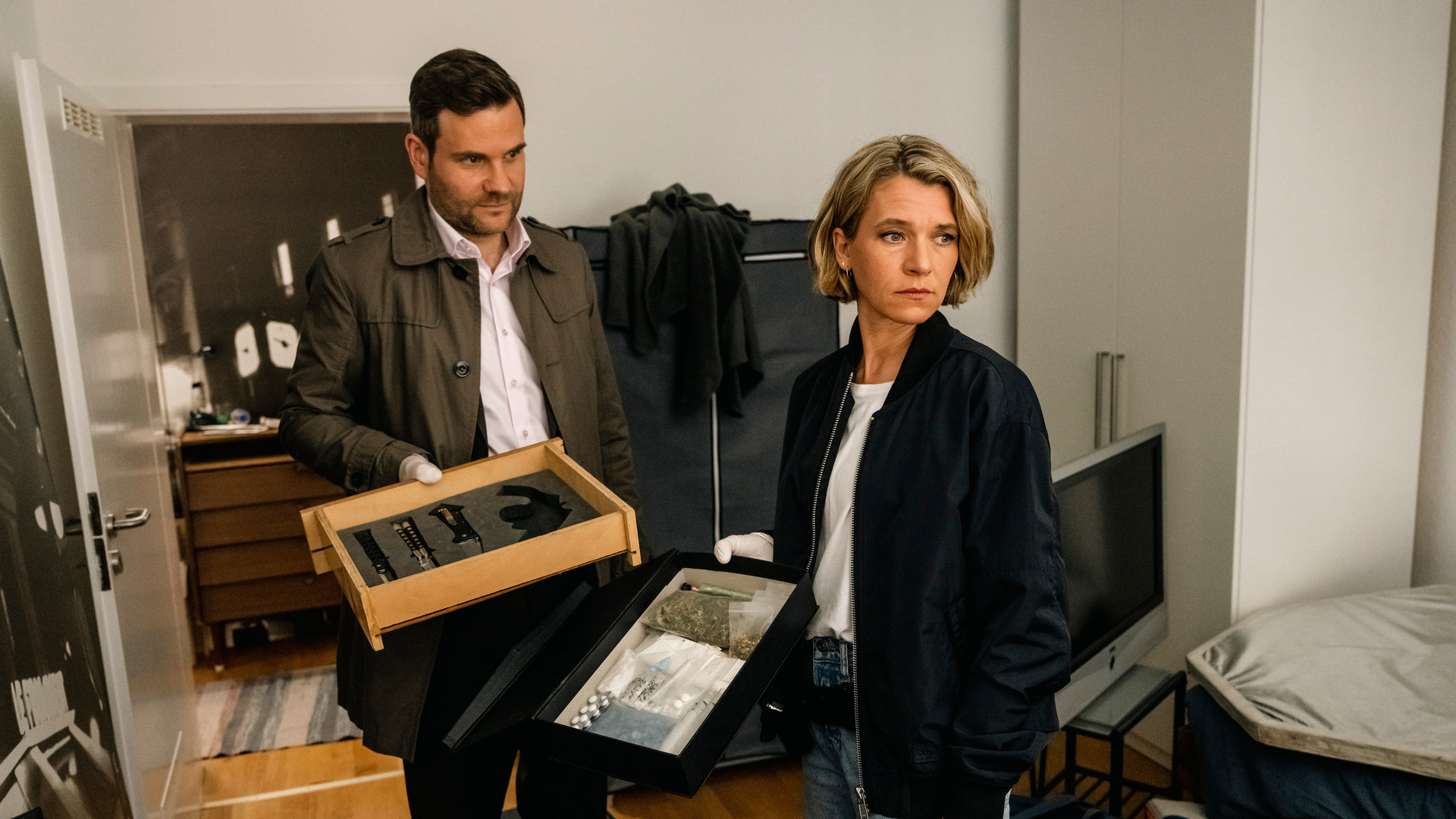 "SOKO Köln - Schuld und Sühne": Vanessa Haas (Kerstin Landsmann) und Jonas Fischer (Lukas Piloty) stehen nebeneinander in einem Wohnzimmer. Vanessa blickt ernst zur Seite, sie hält eine schwarze Schublade mit diversen Kleinigkeiten in der Hand. Jonas sieht seine Kollegin nachdenklich an, er hält eine Schublade mit Messern in der Hand. Im Hintergrund steht eine braune Kommode im Flur der Wohnung, hinter Vanessa steht ein Fernseher auf einem Beistelltisch.