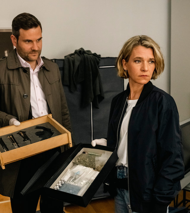 "SOKO Köln - Schuld und Sühne": Vanessa Haas (Kerstin Landsmann) und Jonas Fischer (Lukas Piloty) stehen nebeneinander in einem Wohnzimmer. Vanessa blickt ernst zur Seite, sie hält eine schwarze Schublade mit diversen Kleinigkeiten in der Hand. Jonas sieht seine Kollegin nachdenklich an, er hält eine Schublade mit Messern in der Hand. Im Hintergrund steht eine braune Kommode im Flur der Wohnung, hinter Vanessa steht ein Fernseher auf einem Beistelltisch.