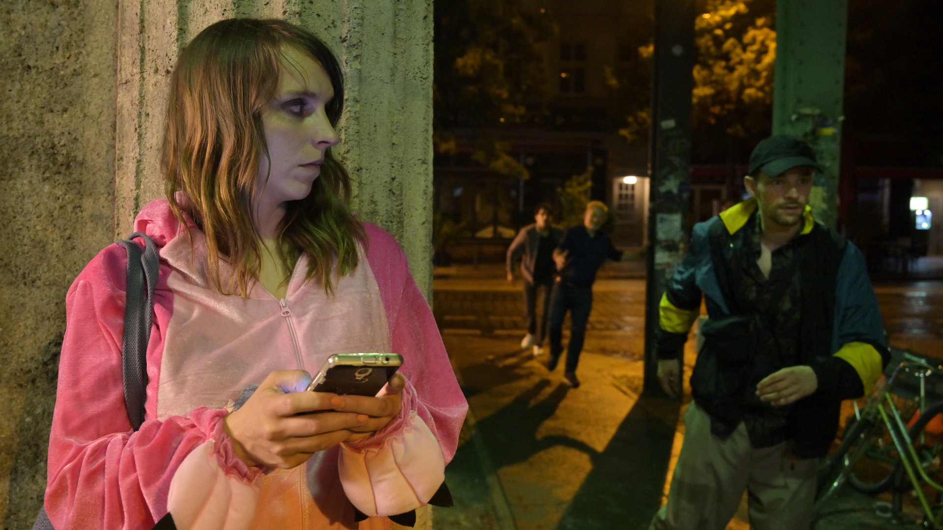 "SOKO Potsdam - Leben lassen": Luna Kunath (Caroline Erikson) steht im rosa Kostüm an einer Straßenecke. Sie hält ein Handy in der Hand und schaut ums Eck, wo ein Mann mit Kappe vorbeirennt.