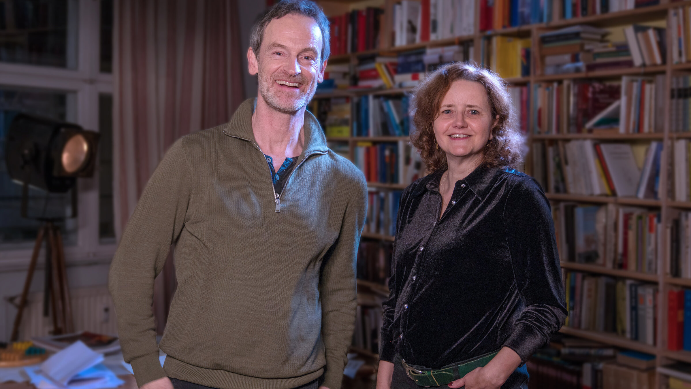 "Bitte um Rückruf - Interview mit einer Schriftstellerin": Schauspieler Jörg Hartmann und Schriftstellerin Julia Schoch stehen nebeneinander vor einer Bücherwand. Beide lächeln in die Kamera.