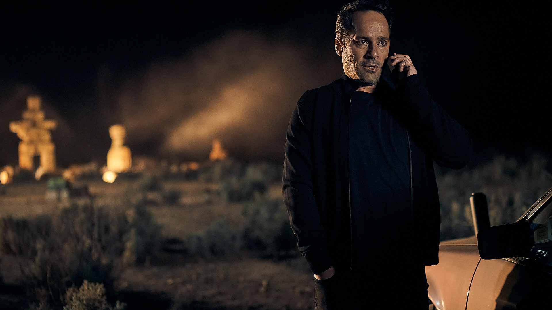 "The Tourist: Precious Memories": Drogenboss Kosta (Alex Dimitriades) steht im nächtlichen Outback neben seinem Auto und telefoniert. Im Hintergrund sieht man die beleuchteten "Steinmänner".