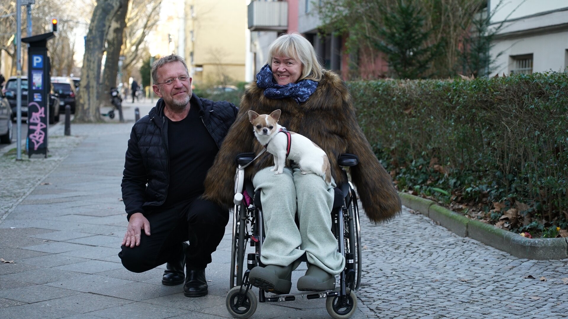"einfach Mensch: Karen Hensel: Selbstbestimmt leben mit Assistenz (1)": Karen Hensel sitzt in ihrem Rollstuhl mit ihrem Hund auf dem Schoß auf einem Bürgersteig. Neben ihr in der Hocke ist ihr Assistenten Hans-Jörg Bahrs. Beide schauen lächelnd in die Kamera.