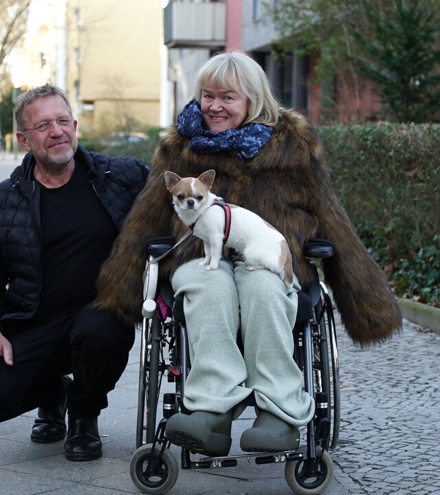 "einfach Mensch: Karen Hensel: Selbstbestimmt leben mit Assistenz (1)": Karen Hensel sitzt in ihrem Rollstuhl mit ihrem Hund auf dem Schoß auf einem Bürgersteig. Neben ihr in der Hocke ist ihr Assistenten Hans-Jörg Bahrs. Beide schauen lächelnd in die Kamera.