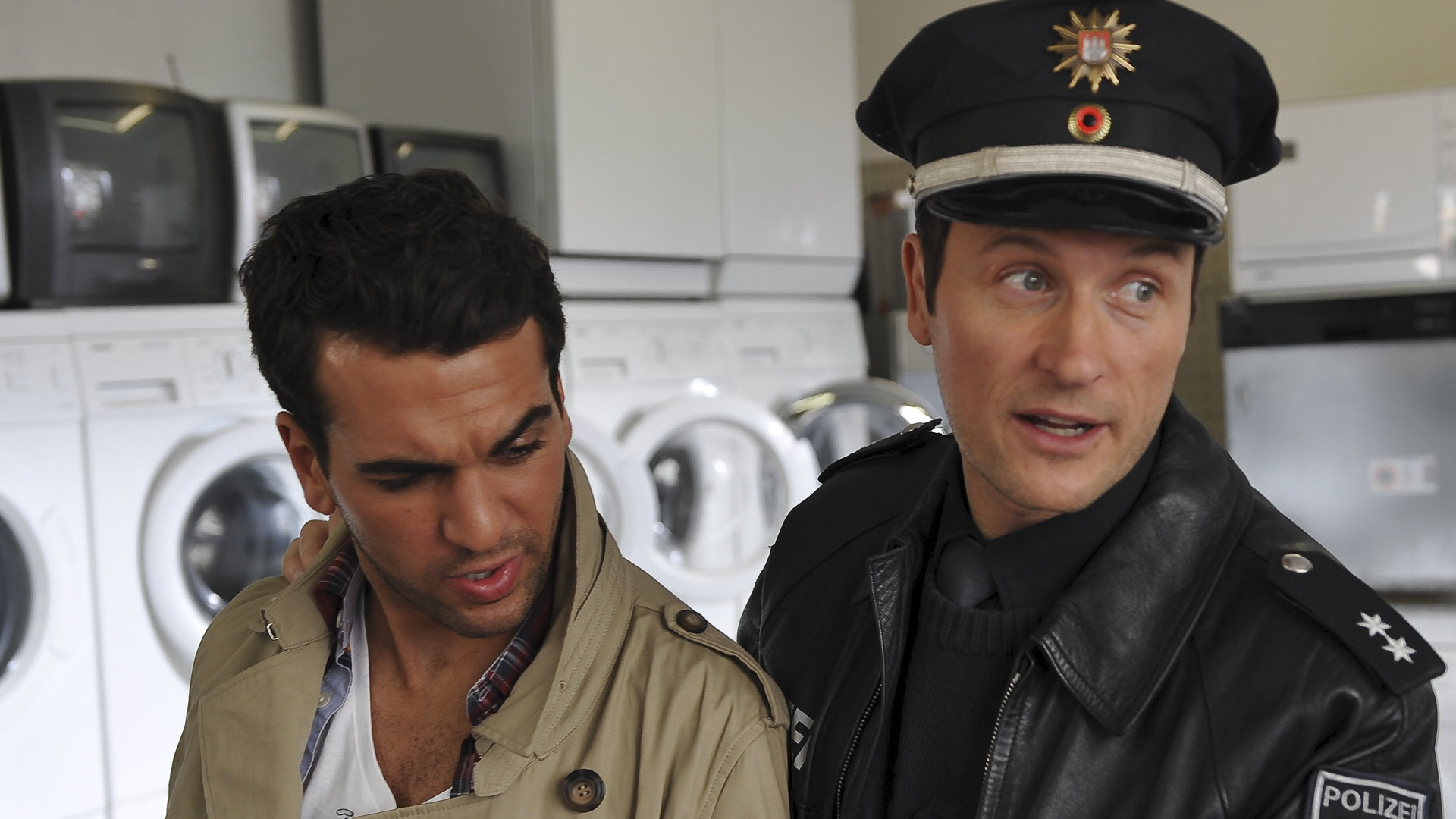 "Notruf Hafenkante - Knock out": Kai (Markus Knüfken) steht neben Hassan (Elias M''Barek) und hält ihn an der Schulter fest. Im Hintergrund stehen Waschmaschinen und Fernsehgeräte.