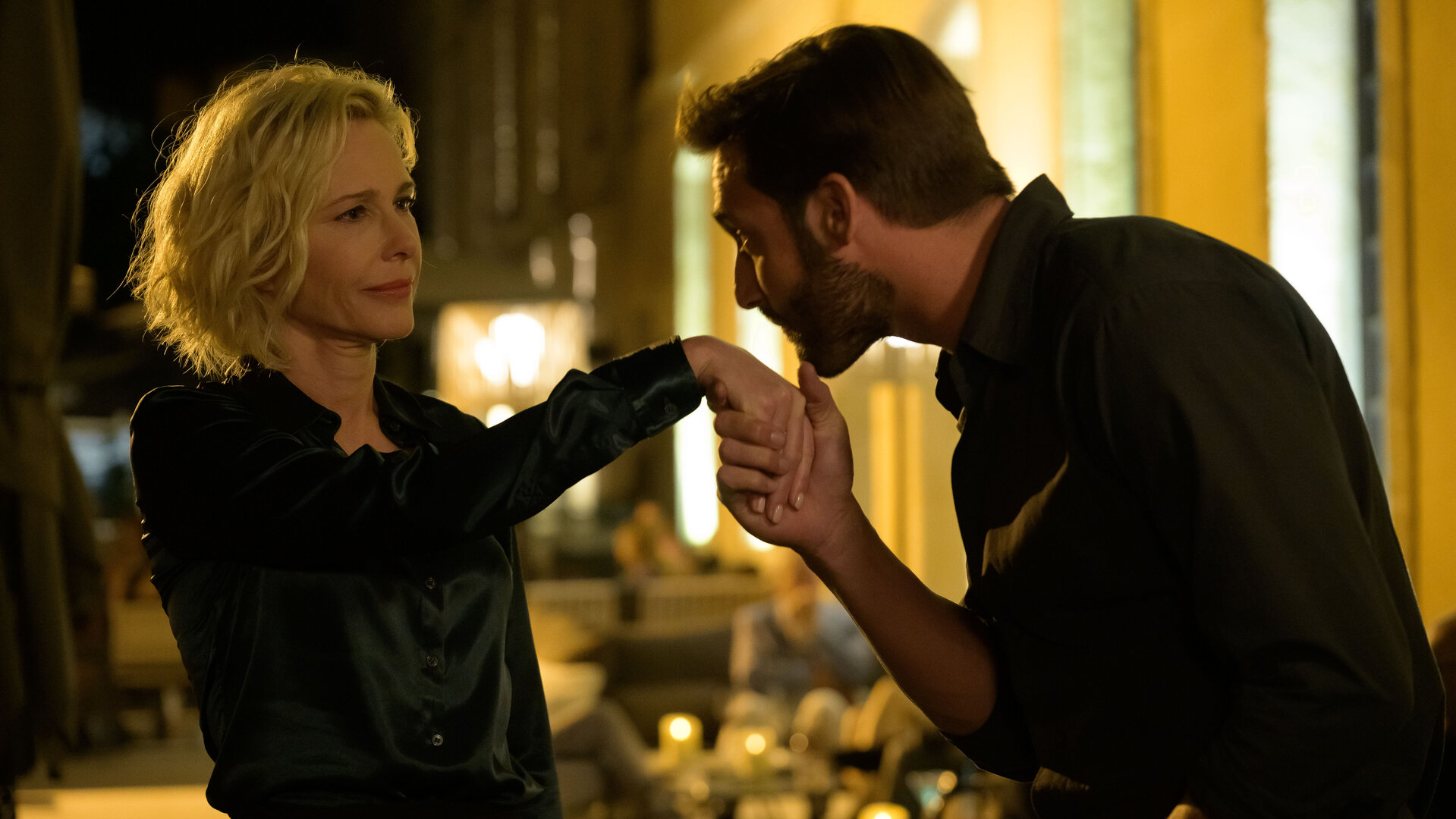 "Escort Boys - Kundenzufriedenheit": Ben (Guillaume Labbé) und Béa (Pascale Arbillot) stehen auf einer Straße. Ben hält Béas Hand und küsst sie, während Béa ihn mit einem leichten Grinsen ansieht. Im Hintergrund stehen Tische, an denen Leute sitzen.