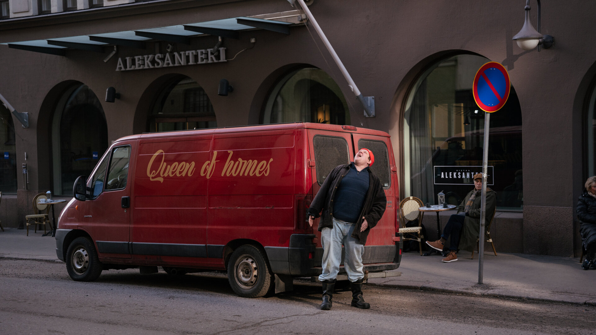 "Queen of Fucking Everything - Die Firma": Vor dem Hotel Aleksanteri in Helsinki ist ein roter Kleinbus mit dem Schriftzug der Firma "Queen of Homes" geparkt. Hannu (Santtu Karvonen) steht an der Rückseite des Fahrzeugs. Er blickt nach oben.