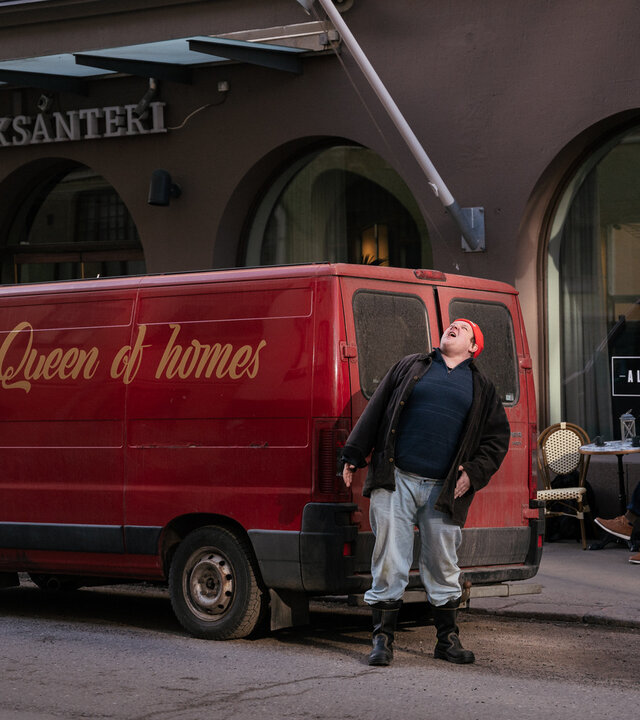 "Queen of Fucking Everything - Die Firma": Vor dem Hotel Aleksanteri in Helsinki ist ein roter Kleinbus mit dem Schriftzug der Firma "Queen of Homes" geparkt. Hannu (Santtu Karvonen) steht an der Rückseite des Fahrzeugs. Er blickt nach oben.