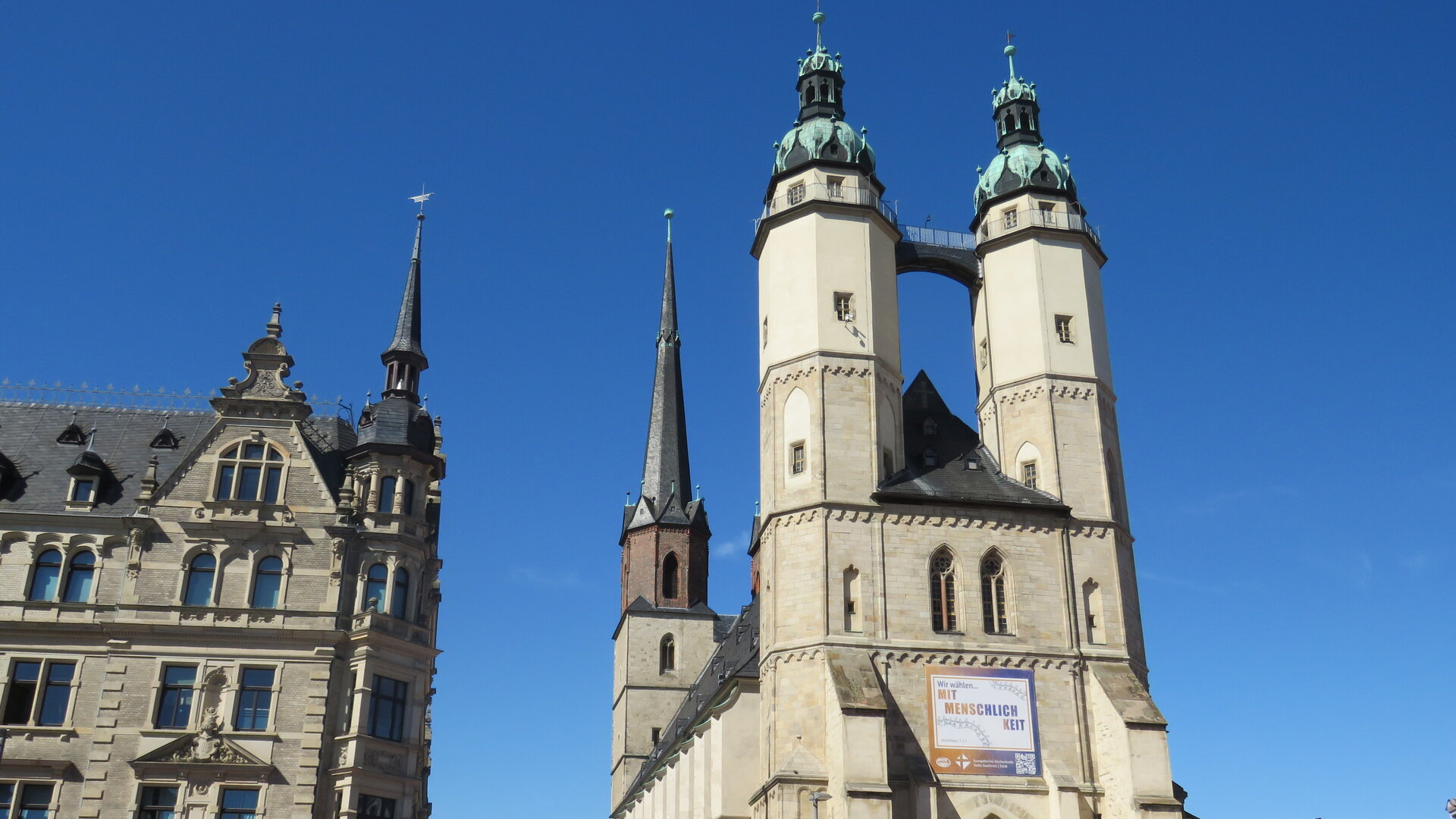"Evangelischer Gottesdienst - Meine Stimme zählt": Untersicht: Die zwei Türme der mittelalterlichen Marktkirche in Halle (Saale) ragen hoch in den blauen Himmel. Daneben befindet sich ein historisches Stadthaus, das mit spitzen Giebeln und ebenfalls mit einem kleinen Türmchen versehen ist.