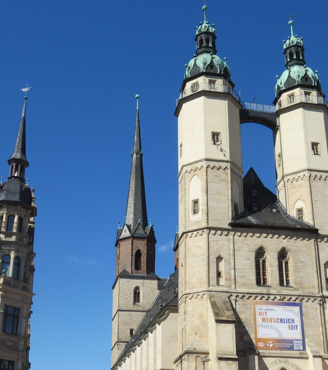 "Evangelischer Gottesdienst - Meine Stimme zählt": Untersicht: Die zwei Türme der mittelalterlichen Marktkirche in Halle (Saale) ragen hoch in den blauen Himmel. Daneben befindet sich ein historisches Stadthaus, das mit spitzen Giebeln und ebenfalls mit einem kleinen Türmchen versehen ist.
