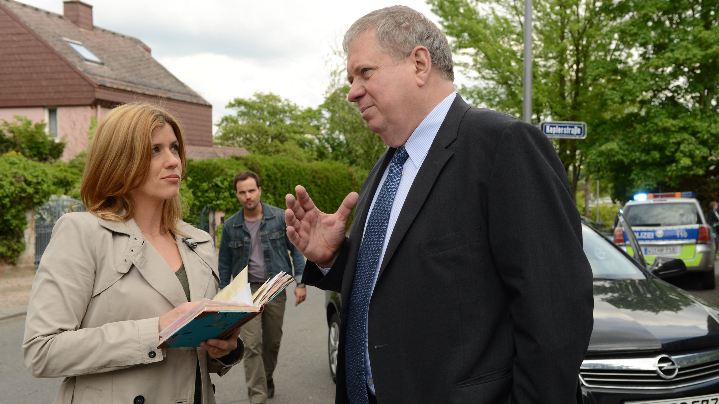 „Der Staatsanwalt - Das Luder“: Kerstin Klar (Fiona Coors), die ein Buch in der Hand hält, und Bernd Reuther (Rainer Hunold) stehen sich auf einer Straße gegenüber. Im Hintergrund erkennt man Christian Schubert (Simon Eckert), der auf die beiden zuläuft.