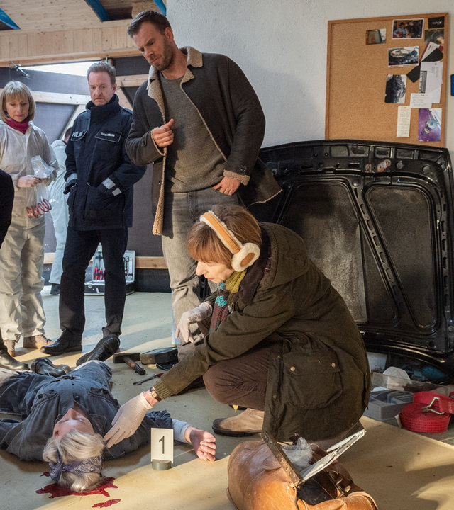 „SOKO Wismar – Benzin im Blut“: Jessy Winter (Josefin Stiller) liegt tot am Boden ihrer Werkstatt. Unter ihrem Kopf ist eine große Blutlache. Um sie herum stehen Karoline Joost (Nike Fuhrmann), Roswitha (Silke Matthias), Timmermann (Mathias Junge) und Lars Pöhlmann (Dominic Boeer). Neben ihrem Kopf kniet Helene Sturbeck (Katharina Blaschke).
