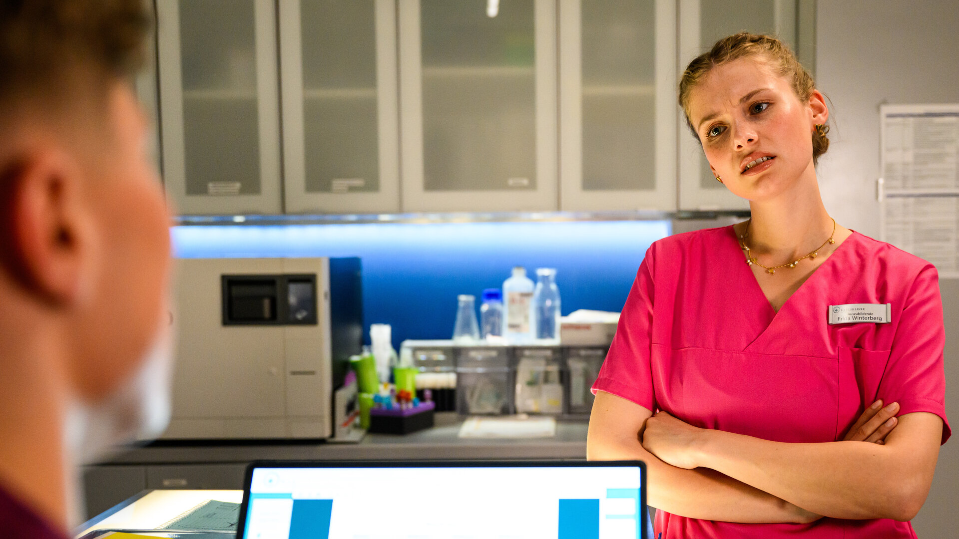 "Bettys Diagnose - Stunde der Wahrheit": Frida (Johanna Hens) steht mit verschränkten Armen Ivo (Oscar Nadermann) gegenüber, der vor einem Laptop sitzt, und schaut ihn an. Im Hintergrund hängt ein Arzneischrank an der Wand und darunter stehen verschiedene Behandlungsutensilien.
