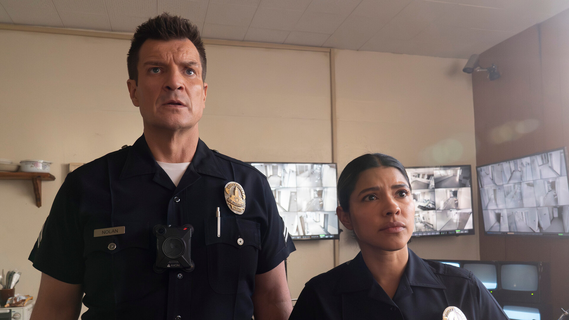 "The Rookie - Westview": Officer John Nolan (Nathan Fillion) und Celina Juarez (Lisseth Chavez) stehen nebeneinander in einem Überwachungsraum. Beide tragen dunkle Polizeiuniformen und blicken fragend nach vorn. Im Hintergrund hängen mehrere Bildschirme mit unterschiedlichen Überwachungsaufnahmen an den Wänden.