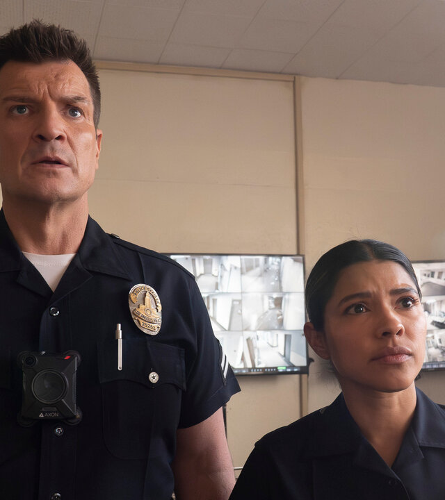 "The Rookie - Westview": Officer John Nolan (Nathan Fillion) und Celina Juarez (Lisseth Chavez) stehen nebeneinander in einem Überwachungsraum. Beide tragen dunkle Polizeiuniformen und blicken fragend nach vorn. Im Hintergrund hängen mehrere Bildschirme mit unterschiedlichen Überwachungsaufnahmen an den Wänden.