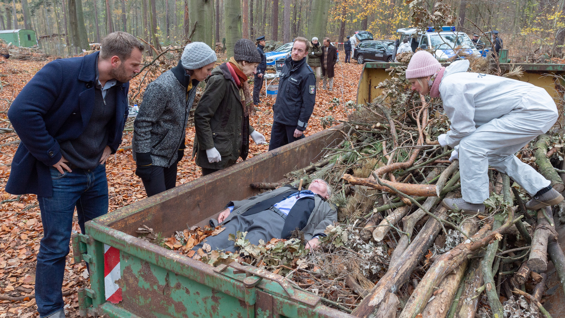 "SOKO Wismar - Schuldbekenntnis": Mitten im Wald steht ein grüner Container mit Waldabfällen. Darin liegt ein Toter im Anzug (Rüdiger Lehmann). Roswitha Prinzler (Silke Matthias) steht auf einem Haufen von Ästen im Container und beugt sich zu dem Toten. Links neben dem Container stehen die Kollegen Pöhlmann (Dominic Boeer), Joost (Nike Fuhrmann), Sturbeck (Katharina Blaschke) und Timmermann (Mathias Junge).