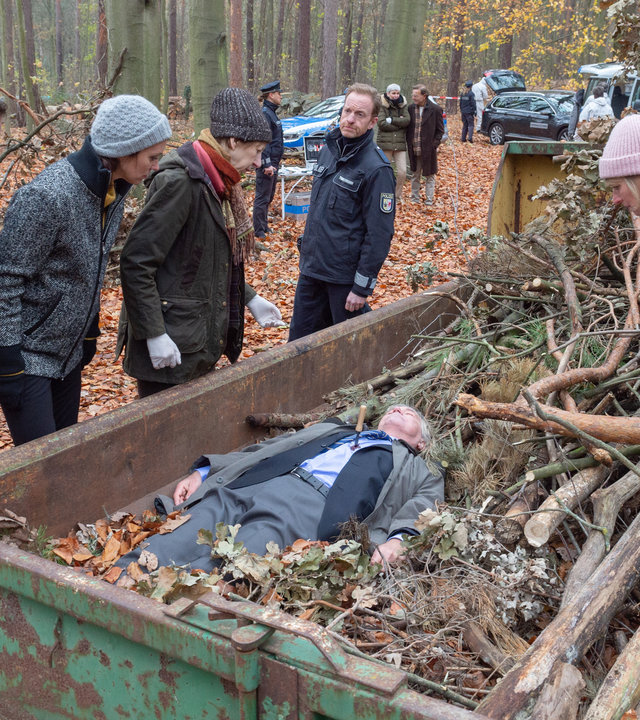 "SOKO Wismar - Schuldbekenntnis": Mitten im Wald steht ein grüner Container mit Waldabfällen. Darin liegt ein Toter im Anzug (Rüdiger Lehmann). Roswitha Prinzler (Silke Matthias) steht auf einem Haufen von Ästen im Container und beugt sich zu dem Toten. Links neben dem Container stehen die Kollegen Pöhlmann (Dominic Boeer), Joost (Nike Fuhrmann), Sturbeck (Katharina Blaschke) und Timmermann (Mathias Junge).