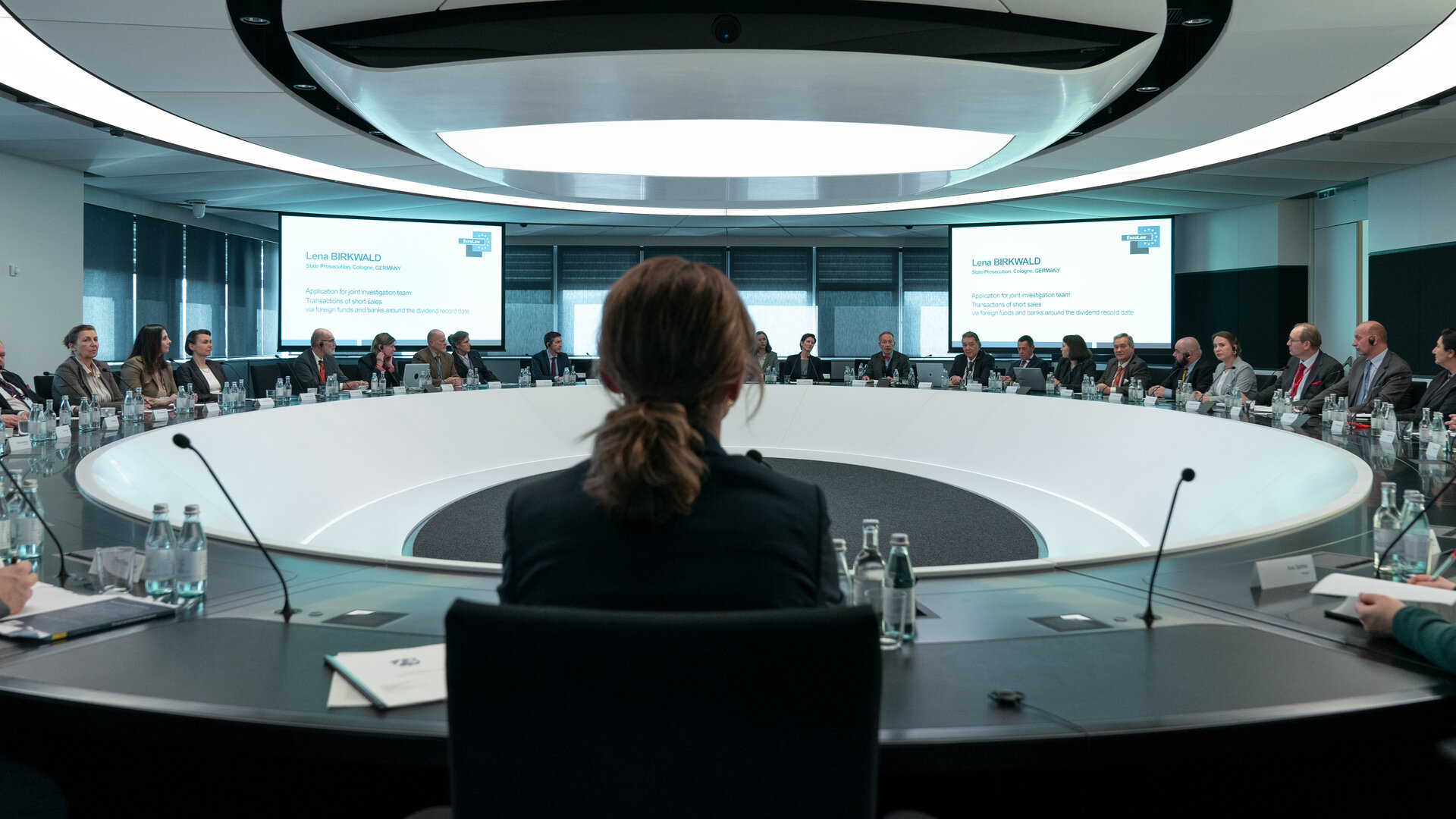 "Die Affäre Cum-Ex - Episode 5": In einem runden Besprechungsraum spricht Lena Birkwald (Lisa Wagner) zu einer Runde von Personen in Anzügen, während im Hintergrund eine Präsentation auf zwei Leinwände projiziert wird.