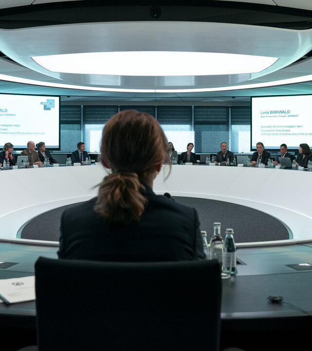 "Die Affäre Cum-Ex - Episode 5": In einem runden Besprechungsraum spricht Lena Birkwald (Lisa Wagner) zu einer Runde von Personen in Anzügen, während im Hintergrund eine Präsentation auf zwei Leinwände projiziert wird.