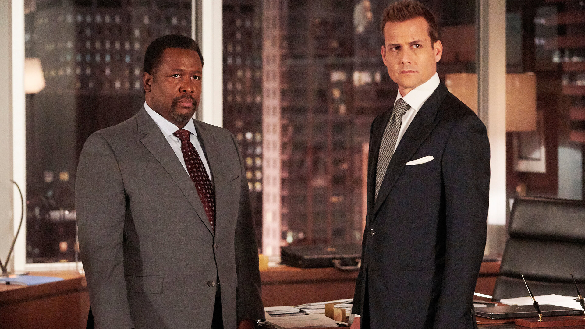 "Suits - Pro-Kopf-Umsatz": Robert (Wendell Pierce, l.) und Harvey (Gabriel Macht)  stehen nebeneinander und schauen beide erwartungsvoll in Richtung Kamera. Sie tragen elegante Anzüge, und im Hintergrund sind die bei Abend erleuchteten Hochhäuser New Yorks zu erkennen.