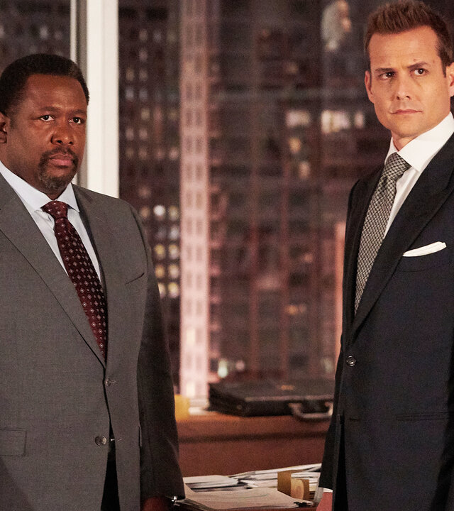 "Suits - Pro-Kopf-Umsatz": Robert (Wendell Pierce, l.) und Harvey (Gabriel Macht)  stehen nebeneinander und schauen beide erwartungsvoll in Richtung Kamera. Sie tragen elegante Anzüge, und im Hintergrund sind die bei Abend erleuchteten Hochhäuser New Yorks zu erkennen.