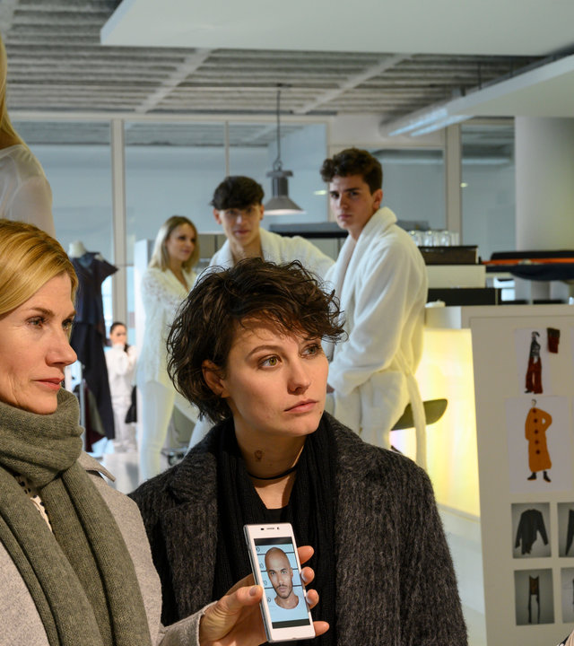 "SOKO Stuttgart – Schön bis in den Tod": Martina Seiffert (Astrid M. Fünderich) und Nele Becker (Nina Siewert) stehen in Wintermänteln gekleidet in den modernen Räumlichkeiten einer Modedesignerin. An den Wänden hängen Modeskizzen. Seiffert hält der Designerin Marie Färber (Fanny Stavjanik) ihr Smartphone entgegen, auf dem ein Foto des Opfers Mick Schuster (Nickolas Weizmann) zu sehen ist. Im Hintergrund stehen Anna Raab (Larissa Marolt) sowie Models (Komparsen) in weißen Bademänteln, die die Befragung beobachten.