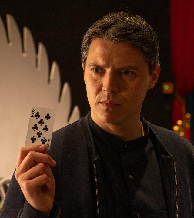 "SOKO Stuttgart - Abrakadabra": Kriminalkommissar Rico Sander (Benjamin Strecker) steht in den Kulissen einer Zaubershow. Mit ernstem Gesichtsausdruck hält er eine Spielkarte in der Hand und zeigt, dass es sich um eine Kreuz Acht handelt.