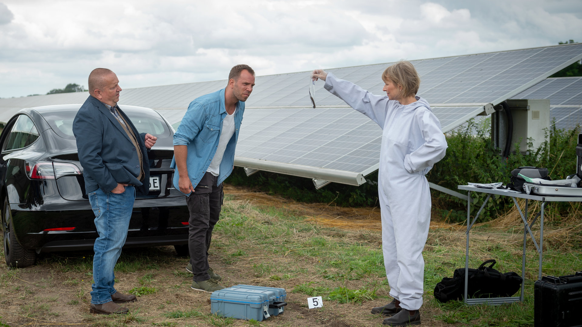 "SOKO Wismar – Tod im Solarfeld": Roswitha Prinzler (Silke Matthias) von der Kriminaltechnik steht im weißen Schutzanzug am Rand eines Solarparks und hält ein Beweismittel in die Höhe. Pöhlmann (Dominic Boeer) und Reuter (Udo Kroschwald) betrachten es sich gemeinsam.