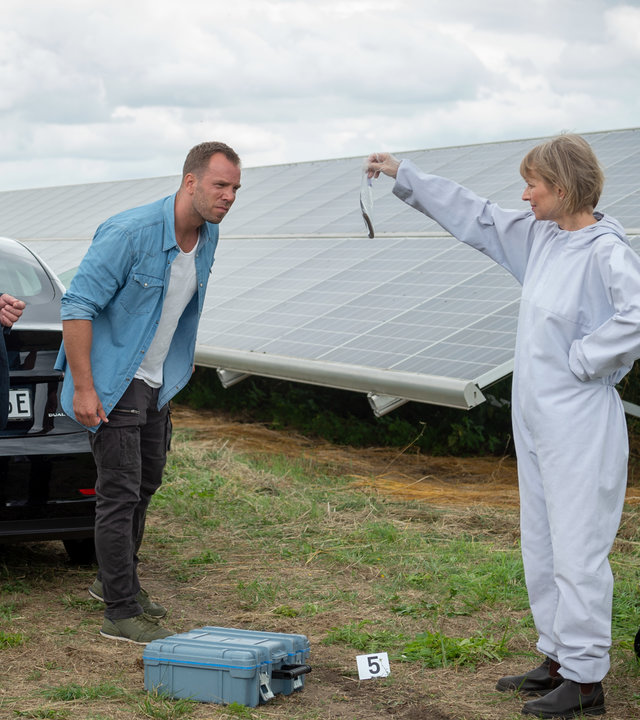 "SOKO Wismar – Tod im Solarfeld": Roswitha Prinzler (Silke Matthias) von der Kriminaltechnik steht im weißen Schutzanzug am Rand eines Solarparks und hält ein Beweismittel in die Höhe. Pöhlmann (Dominic Boeer) und Reuter (Udo Kroschwald) betrachten es sich gemeinsam.