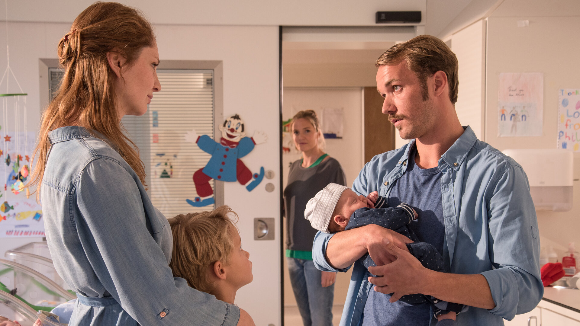 "Lena Lorenz - Eindeutig uneindeutig (2)": In einem Kinderkrankenzimmer stehen sich Nina (Daniela Schulz) und Thomas Cramer (Max Woelky) gegenüber. Thomas hält ein Baby im Arm. Aaron Cramer (Simon Tiefenbacher), ein etwa achtjähriger Junge, schmiegt sich an seine Mutter. Im Hintergrund steht Lena Lorenz (Patricia Aulitzky) in der Tür und beobachtet die Familie mit einem zufriedenen Gesichtsausdruck.