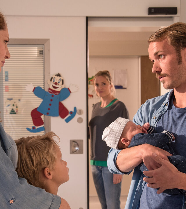 "Lena Lorenz - Eindeutig uneindeutig (2)": In einem Kinderkrankenzimmer stehen sich Nina (Daniela Schulz) und Thomas Cramer (Max Woelky) gegenüber. Thomas hält ein Baby im Arm. Aaron Cramer (Simon Tiefenbacher), ein etwa achtjähriger Junge, schmiegt sich an seine Mutter. Im Hintergrund steht Lena Lorenz (Patricia Aulitzky) in der Tür und beobachtet die Familie mit einem zufriedenen Gesichtsausdruck.