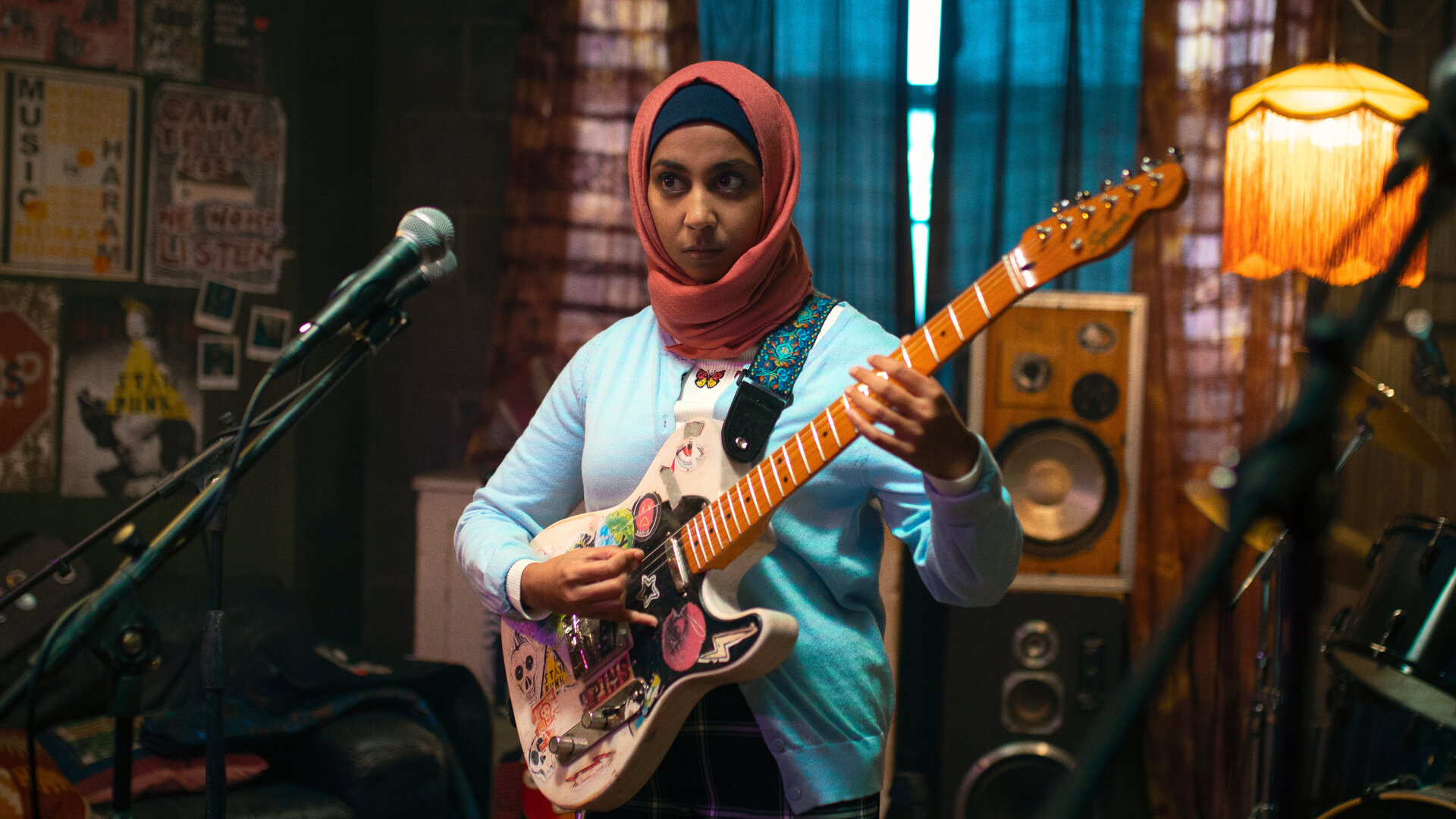 "We Are Lady Parts - Potenzieller zukünftiger Ehemann": Amina (Anjana Vasan) steht mit einer bunt beklebten E-Gitarre in einem Proberaum und greift konzentriert in die Saiten. Vor ihr befinden sich Mikrofone, im Hintergrund sind Lautsprecher, eine Stehlampe und ein Schlagzeug mit dem Schriftzug "Lady Parts" zu sehen.