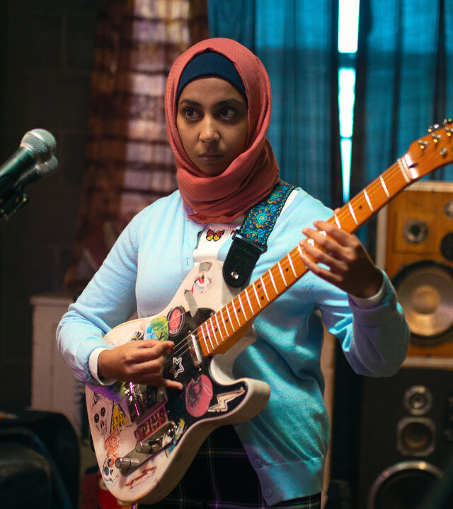 "We Are Lady Parts - Potenzieller zukünftiger Ehemann": Amina (Anjana Vasan) steht mit einer bunt beklebten E-Gitarre in einem Proberaum und greift konzentriert in die Saiten. Vor ihr befinden sich Mikrofone, im Hintergrund sind Lautsprecher, eine Stehlampe und ein Schlagzeug mit dem Schriftzug "Lady Parts" zu sehen.