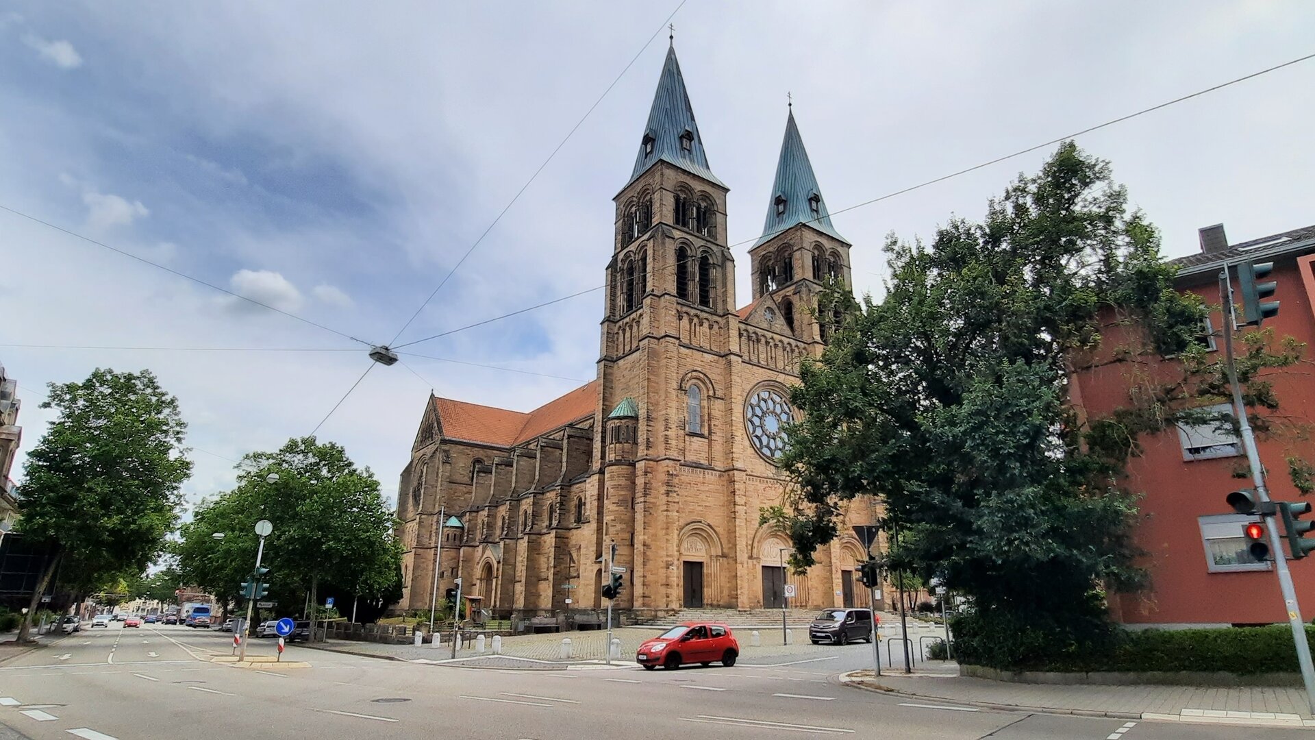 "Katholischer Gottesdienst - Beseelte Orte": Außenansicht der Marienkirche in Landau i. d. Pfalz: Die Kirche steht an einer Kreuzung, zwei Türme ragen in den bewölkten Himmel.