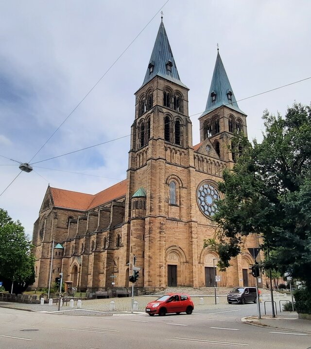 "Katholischer Gottesdienst - Beseelte Orte": Außenansicht der Marienkirche in Landau i. d. Pfalz: Die Kirche steht an einer Kreuzung, zwei Türme ragen in den bewölkten Himmel.