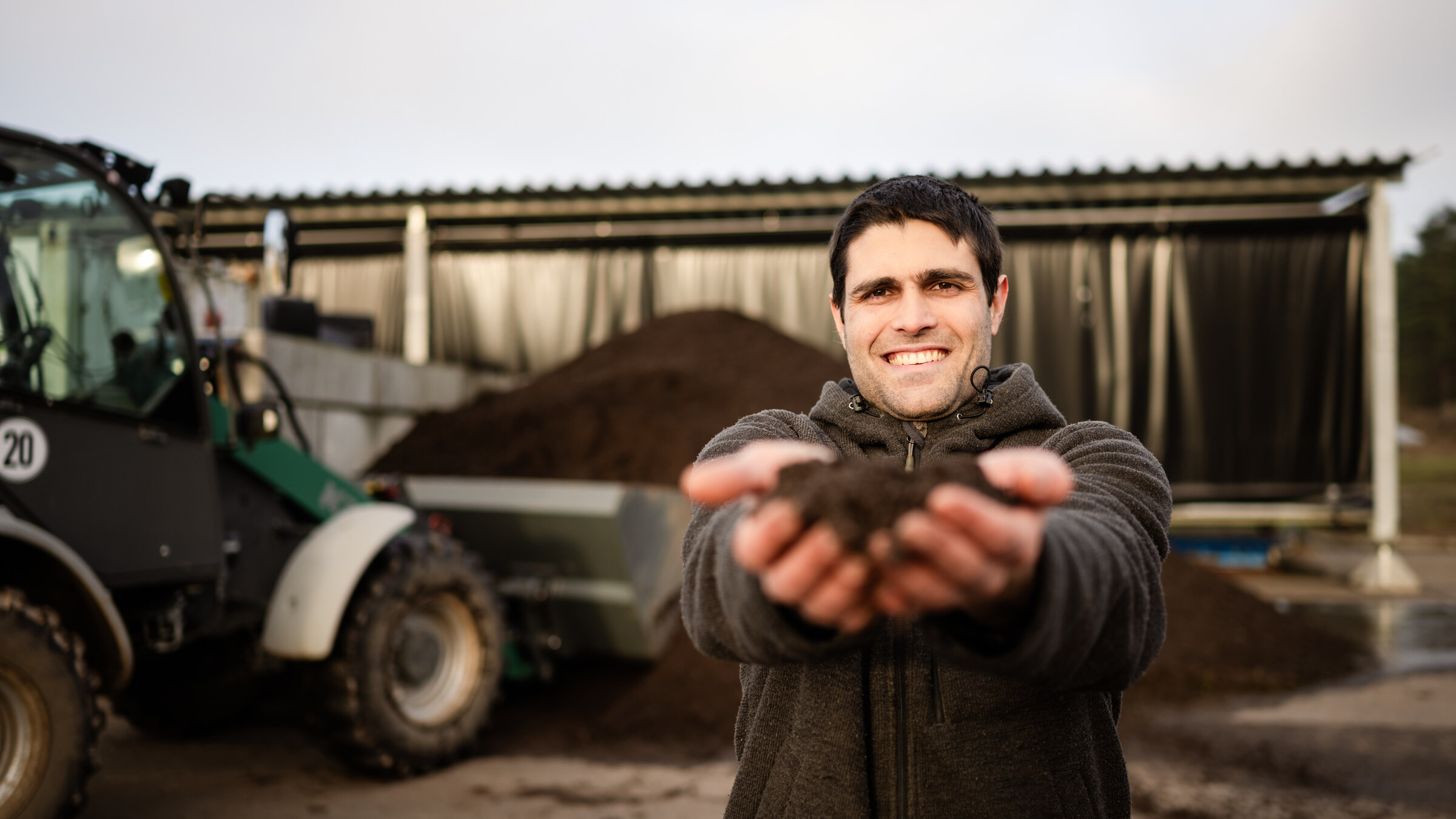 "Kreative Köpfe, große Träume - Nachhaltig wirtschaften": Florian steht vor einer Halle neben einem Traktor. Er hat die Arme ausgestreckt und hält in beiden Händen seinen Dünger. Er schaut lächelnd in die Kamera.