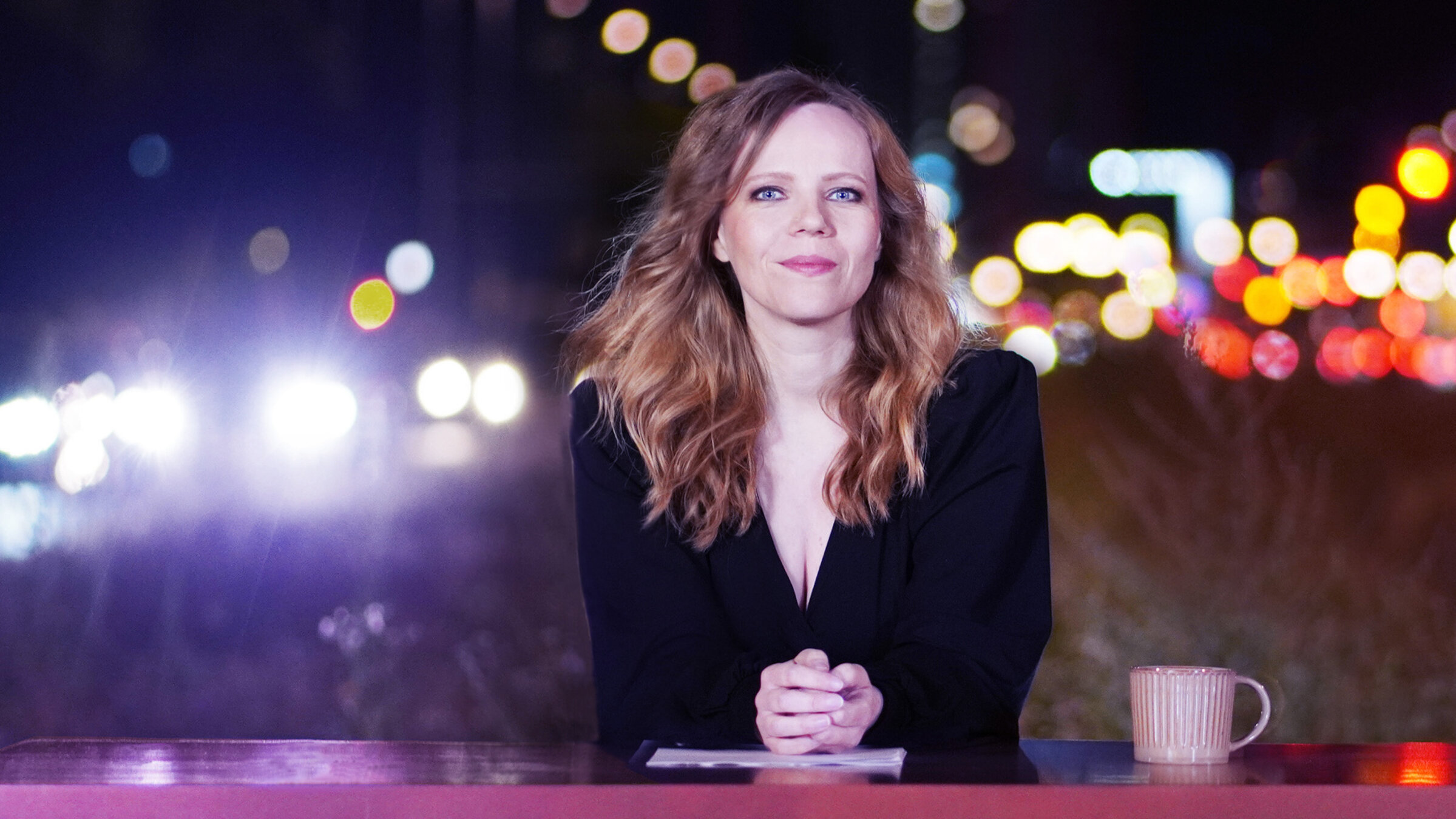 "Bosetti Late Night": Sarah Bosetti sitzt hinter einem pinkfarbenen Tresen. Sie schaut freundlich in die Kamera. Neben Ihr steht eine rosafarbene Kaffeetasse.