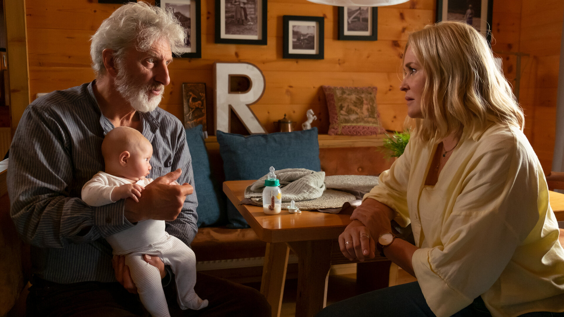 "Lena Lorenz – Wahlverwandtschaft": In einem rustikalen Esszimmer mit Holzvertäfelung und Familienfotos an den Wänden sitzen sich Ansgar Lenz (Rufus Beck) und Lena Lorenz (Judith Hoersch) gegenüber. Lena schaut Ansgar, der ein Baby auf dem Schoß hält,  mitfühlend an.