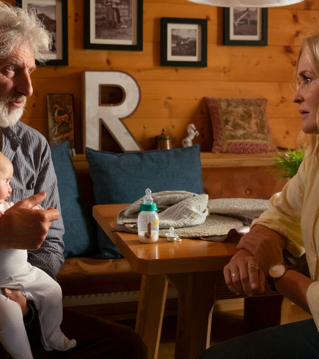 "Lena Lorenz – Wahlverwandtschaft": In einem rustikalen Esszimmer mit Holzvertäfelung und Familienfotos an den Wänden sitzen sich Ansgar Lenz (Rufus Beck) und Lena Lorenz (Judith Hoersch) gegenüber. Lena schaut Ansgar, der ein Baby auf dem Schoß hält,  mitfühlend an.