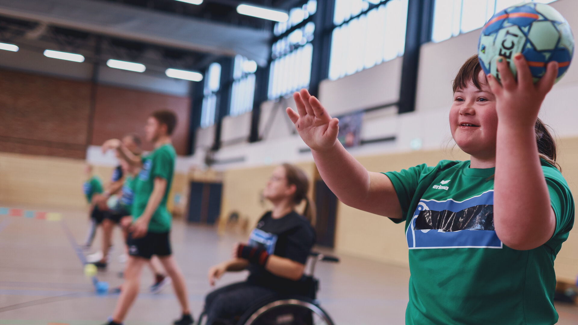 "Aktion Mensch - Glückszahlen der Woche": In einer Sporthalle trainieren mehrere Personen Handball. Im Vordergrund steht ein junges Mädchen, die einen Handball in Wurfposition hält. Hinter ihr sitzt eine Person im Rollstuhl.