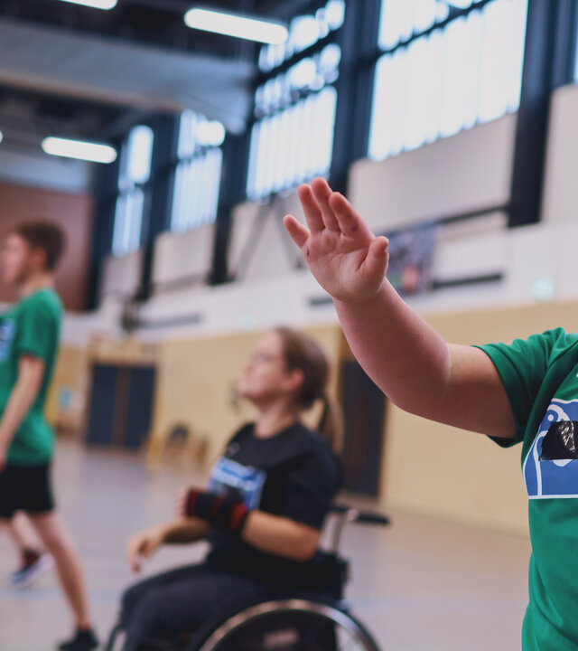 "Aktion Mensch - Glückszahlen der Woche": In einer Sporthalle trainieren mehrere Personen Handball. Im Vordergrund steht ein junges Mädchen, die einen Handball in Wurfposition hält. Hinter ihr sitzt eine Person im Rollstuhl.