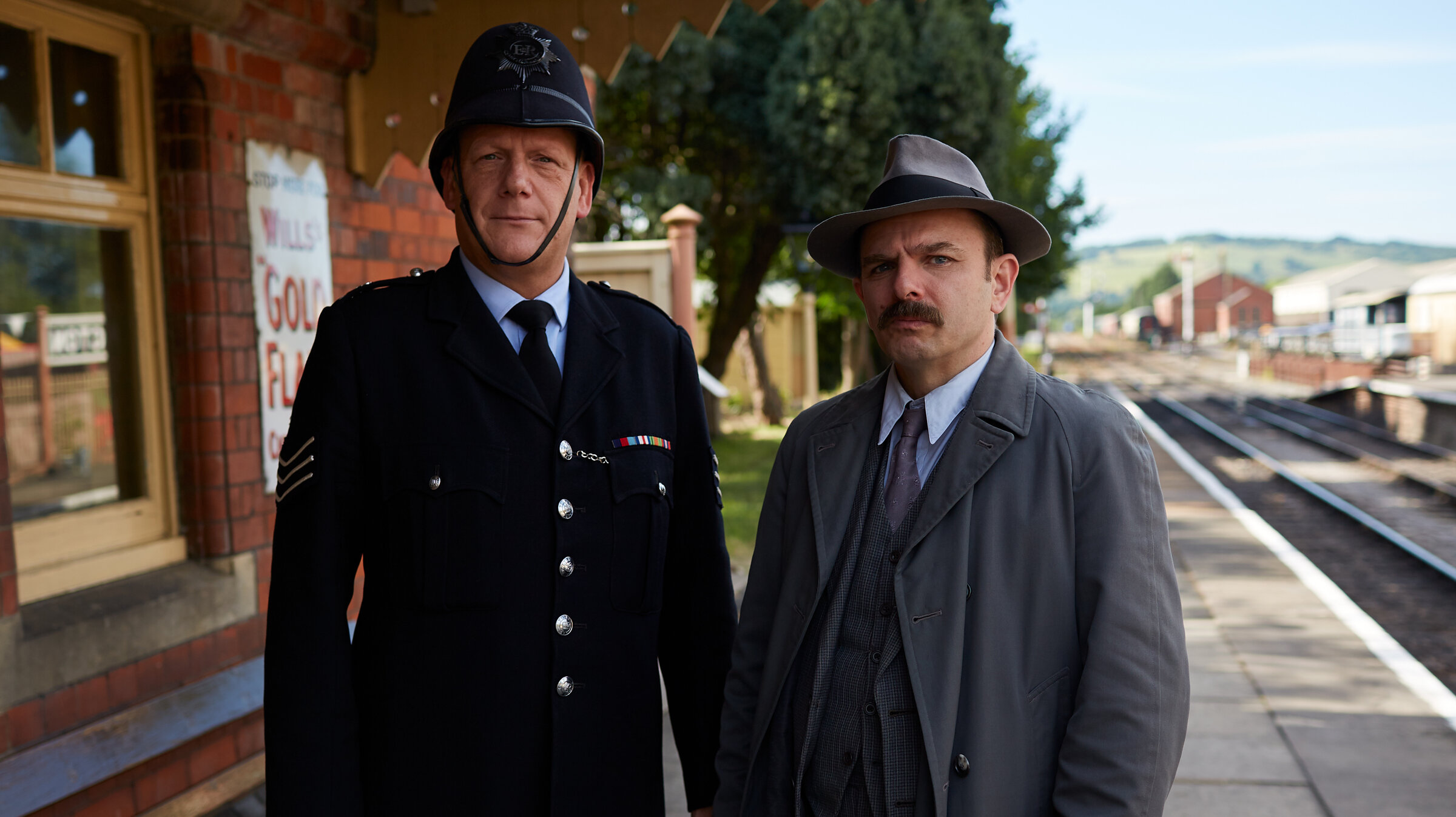 "Father Brown - Der Drache von Kembleford": Sergeant Goodfellow (John Burton) und Inspector Mallory (Jack Deam) stehen auf dem Bahnsteig. Beide blicken konzentriert in Richtung Kamera.