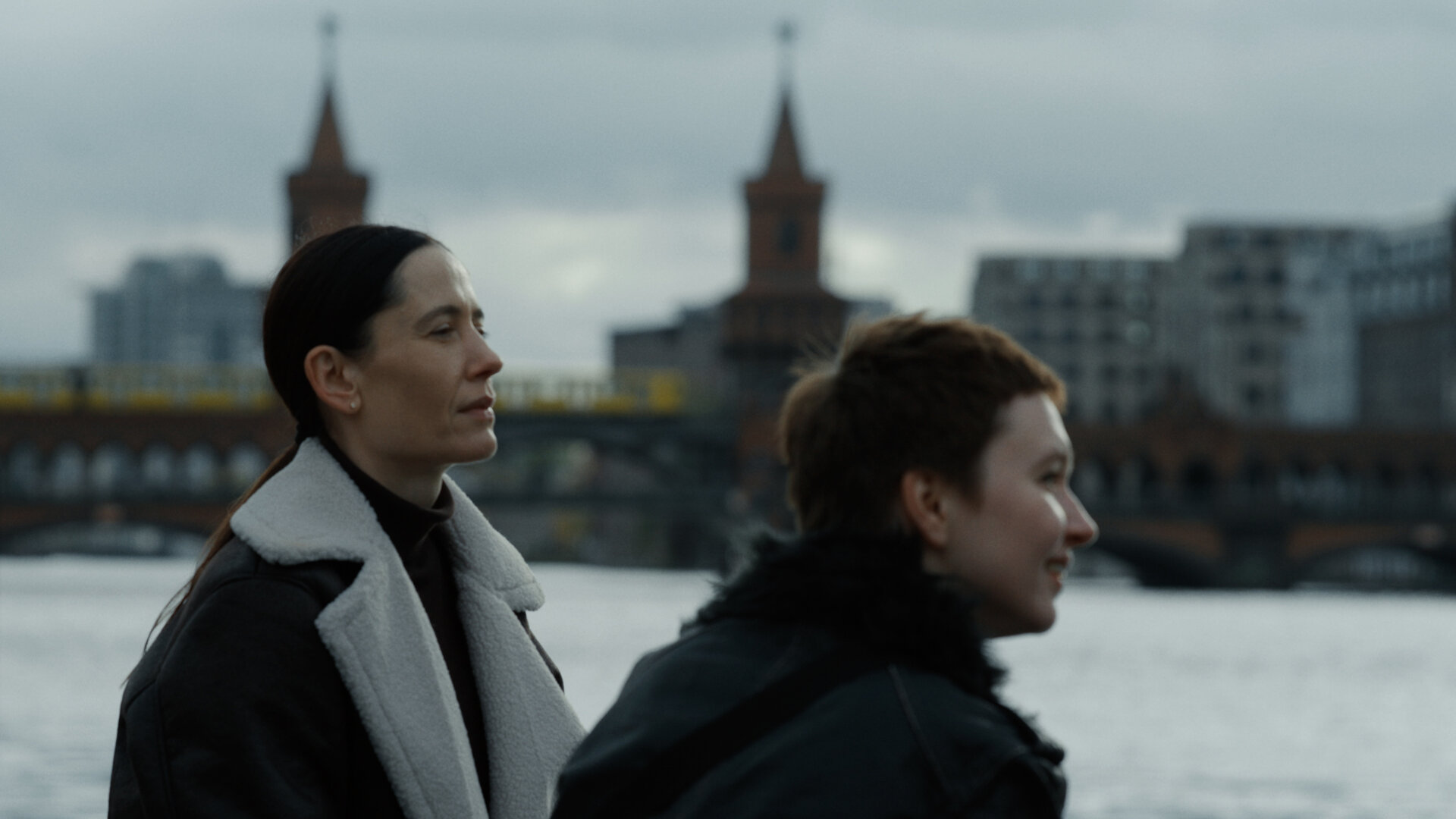 "In Her Car: Die verwunschene Flöte": Lydia (Anastasia Karpenko) und Dasha (Kateryna Hryhorenko) stehen nebeneinander und blicken auf die Spree in Berlin. Im Hintergrund sind die Türme der Oberbaumbrücke erkennbar.