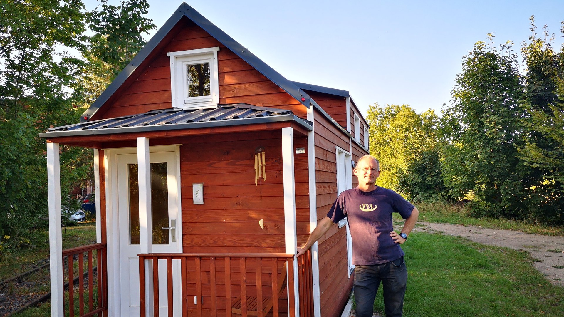 "Terra Xpress: Mietest Du noch oder baust Du schon?": Oliver Victor steht vor einem seiner braunen Tiny-Houses und lehnt sich an den Zaun der kleinen Veranda.
